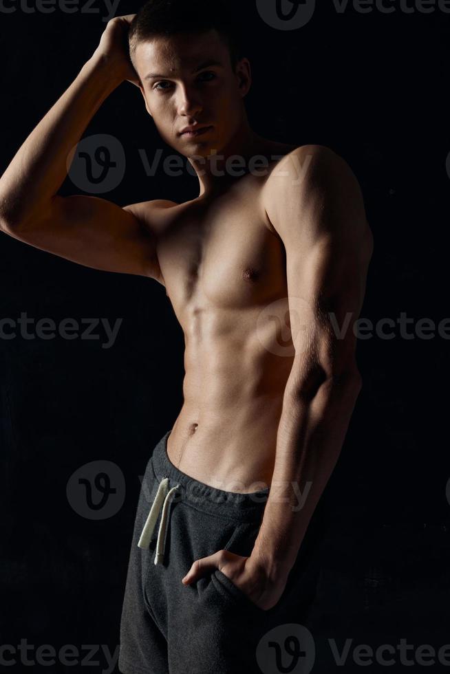 Athlet Bodybuilder mit Hand in der Nähe von das Gesicht und im grau Hose auf ein schwarz Hintergrund foto