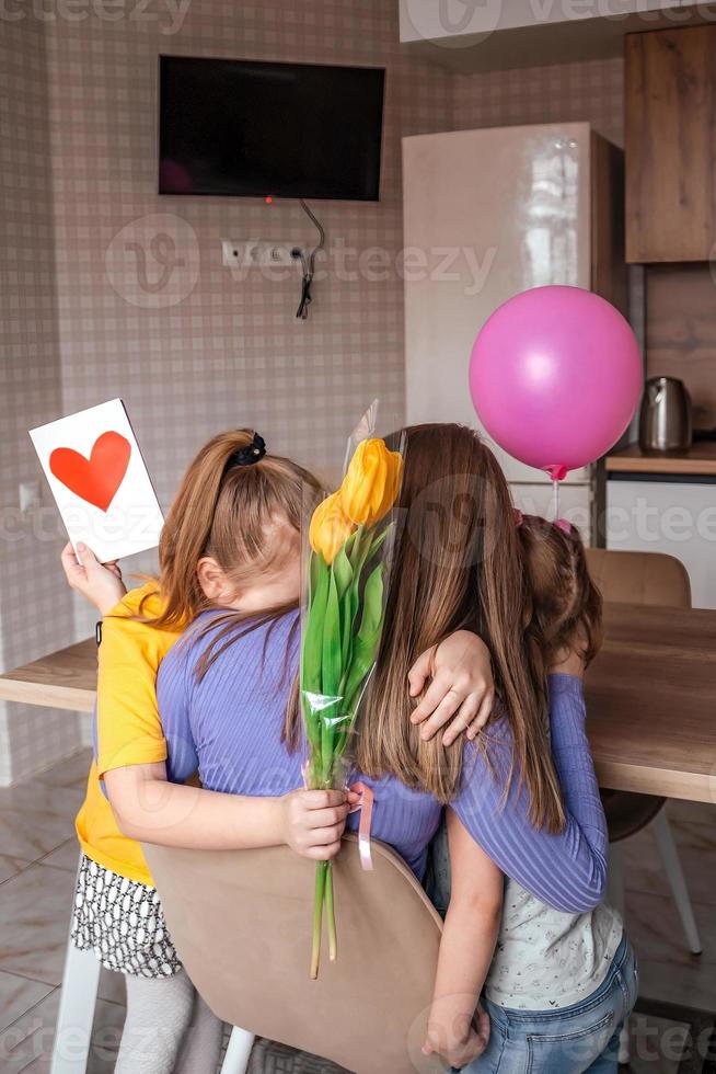 Töchter gratulieren ihr Mama auf Mutter Tag, ein Karte mit ein Herz, Blumen und ein Ballon beim Zuhause im das Küche, Sie alle Umarmung. Kinder Überraschung ihr Mutter zum das Urlaub. foto