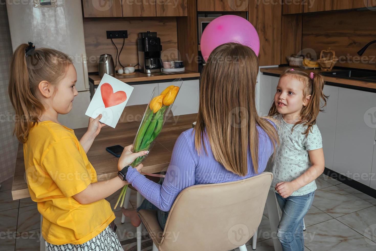 Töchter gratulieren ihr Mama auf Mutter Tag, ein Karte mit ein Herz, Blumen und ein Ballon beim Zuhause im das Küche. Kinder Überraschung ihr Mutter zum das Urlaub. foto