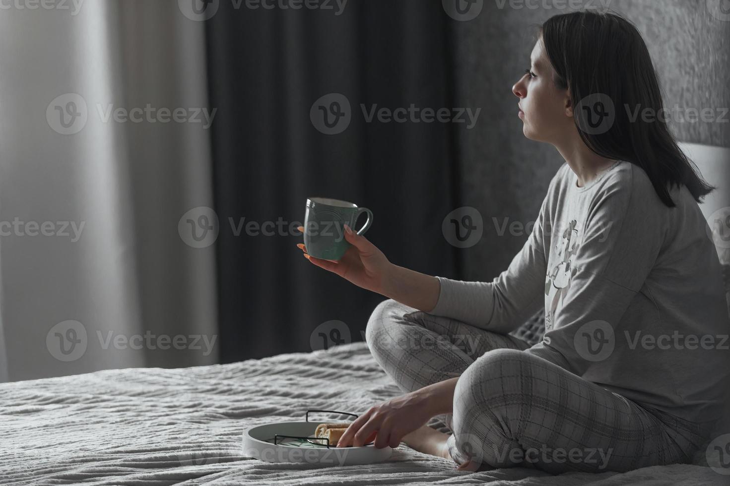 ein schön jung Mädchen im Pyjama sitzt allein auf das Bett im das Morgen im ihr Schlafzimmer, im ihr Hände ist ein Becher von Tee, sie sieht aus aus das Fenster foto