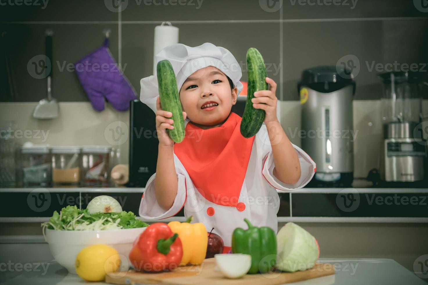 süß asiatisch Mädchen tragen Koch Uniform mit ein Menge von vegatable auf das Tabelle im das Küche Zimmer, machen Essen zum Essen Abendessen, lustig Zeit zum Kinder foto