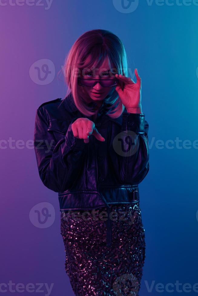 verwirrt sexy schön blond Frau im Leder Jacke prickelnd Kleid modisch Sonnenbrille Punkt Finger beim Kamera posieren isoliert im Blau Rosa Farbe Licht Studio Hintergrund. Neon- Party Cyberpunk Konzept foto