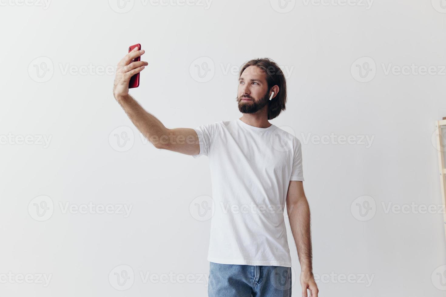 ein männlich Blogger Videoaufnahmen selbst auf seine Telefon und chatten mit Menschen online mit ein Lächeln im ein Weiß T-Shirt gegen ein Weiß Mauer foto