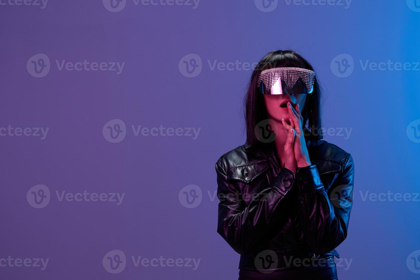 erschrocken schockiert genial brünett Frau im Leder Jacke modisch spiegelnd Sonnenbrille falten Hände in der Nähe von Gesicht posieren isoliert im Blau violett Farbe Licht Hintergrund. Neon- Party Cyberpunk Konzept. Kopieren Raum foto