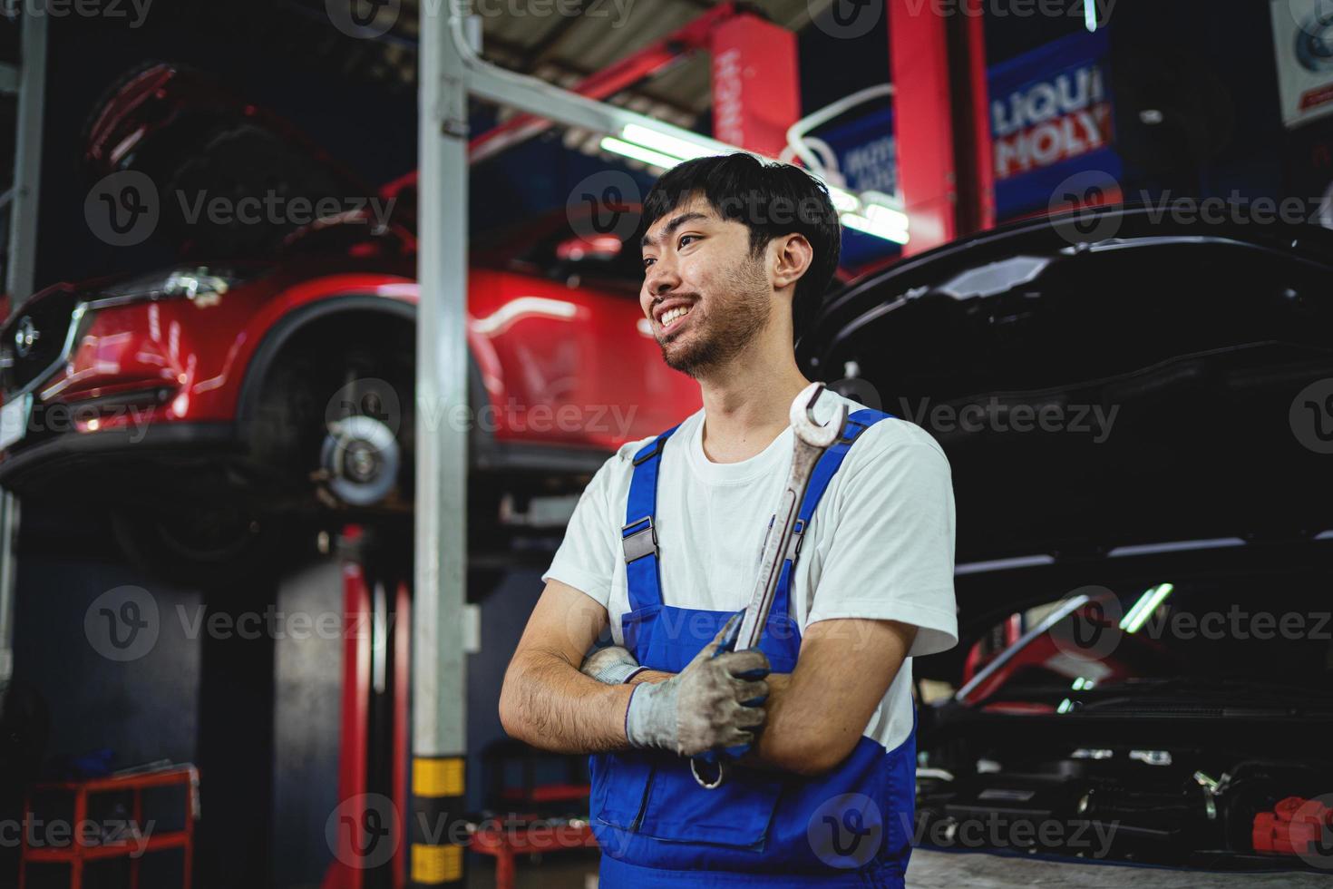 Porträt von lächelnd asiatisch männlich Handwerker halten mit gekreuzten Beinen Schlüssel beim Garage Fachmann Reparatur und Wartung. Auto Inspektion Bedienung foto
