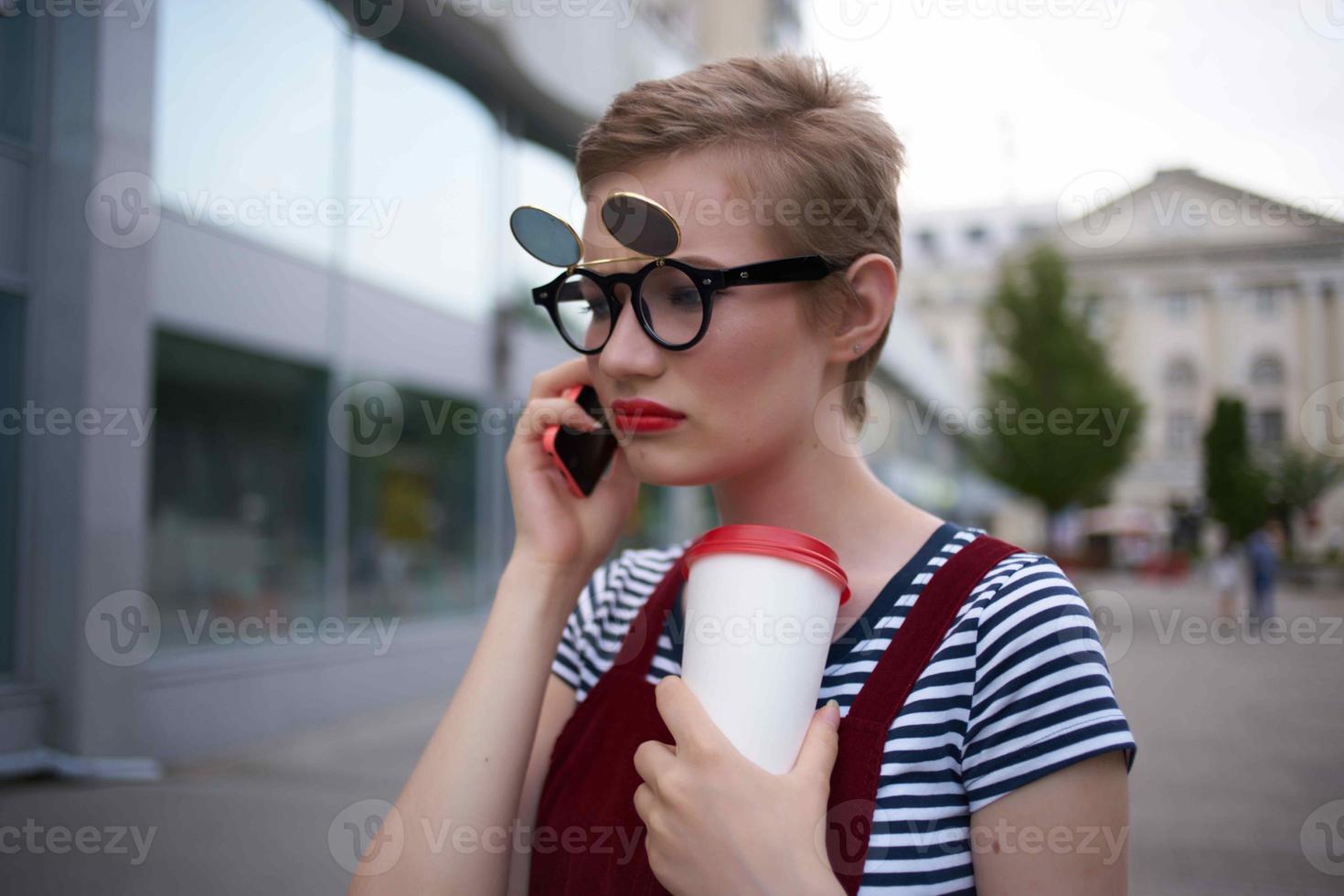 Frau mit Brille auf das Straße reden auf das Telefon im ein Glas mit ein trinken foto