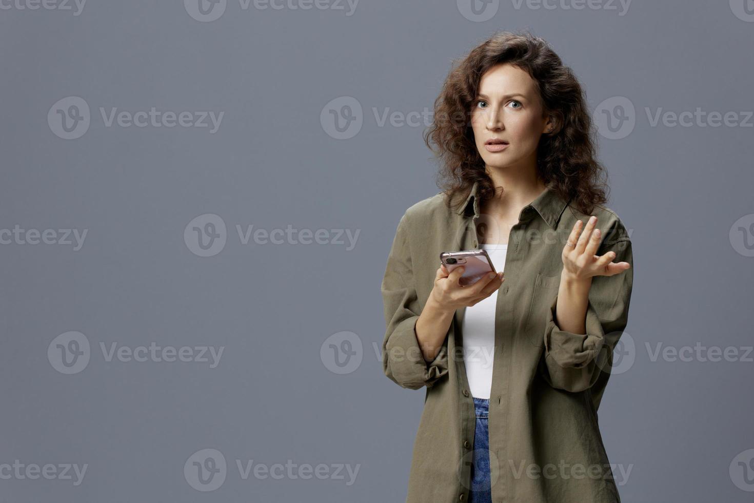 verwirrt unzufrieden wütend lockig schön Frau im beiläufig khaki Grün Hemd erhalten Fälschung Botschaft von Betrug mit Telefon posieren isoliert auf grau Blau Hintergrund. Sozial Medien Influencer Konzept. Kopieren Raum foto