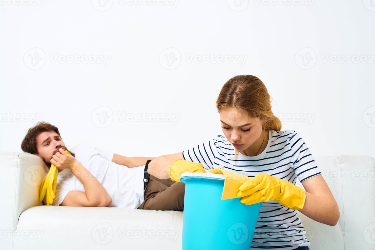 Frau mit ein Eimer im Gummi Handschuhe beim Zuhause Mann Lügen auf das Couch Reinigung foto