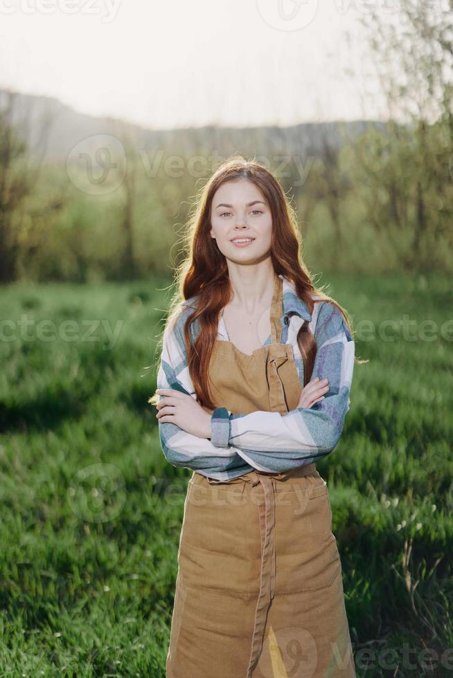 ein Farmer Frau im ihr Arbeit Kleidung, Plaid Hemd und Schürze, steht im das Feld auf das Grün Gras und lächelt im das Rahmen Sonne foto