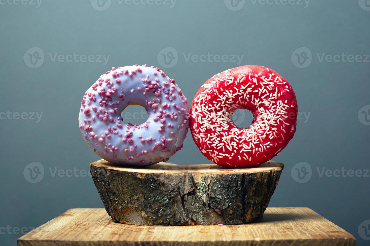 zwei runde glasierte Donuts, bestreut mit süßem Zuckergussrot und Purpur auf einem Holzsockel auf grauem Hintergrund foto