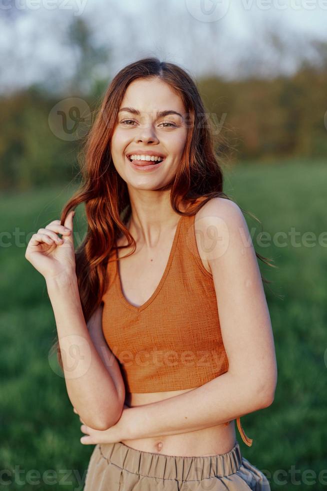 komisch jung rothaarig Frau im ein Orange oben lächelnd gegen das Grün Gras von Natur im das Sonnenuntergang Licht foto