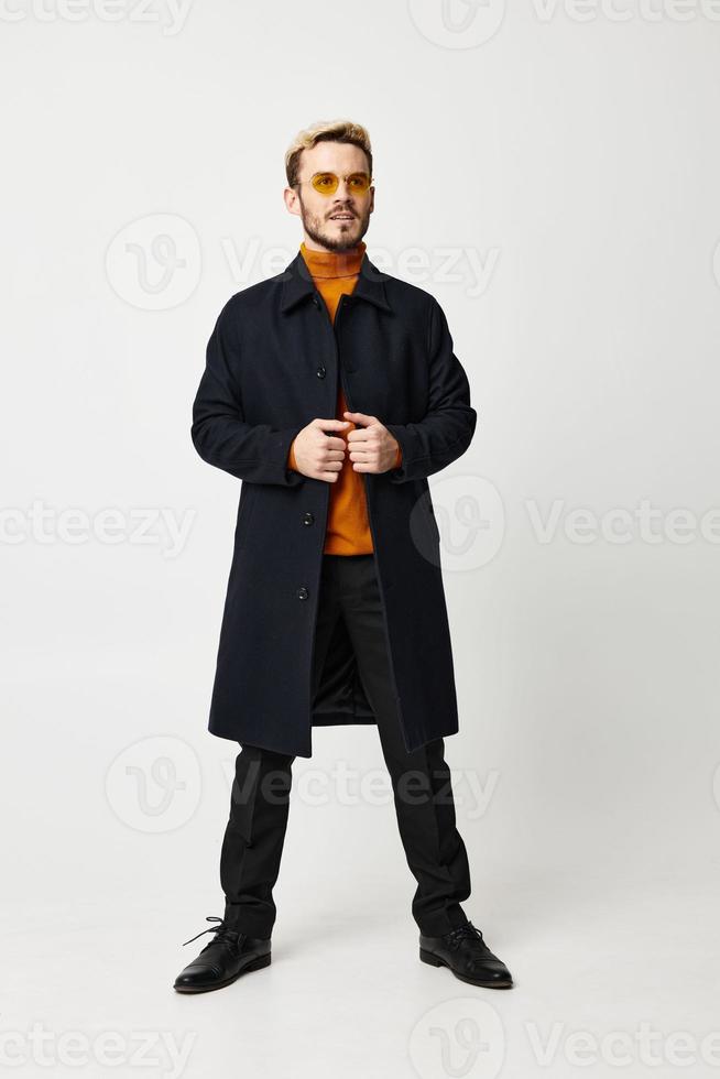 modisch Mann im ein dunkel Mantel Verbreitung seine Beine Schulterbreite ein Teil auf ein Licht Hintergrund und ein Orange Sweatshirt Modell- foto