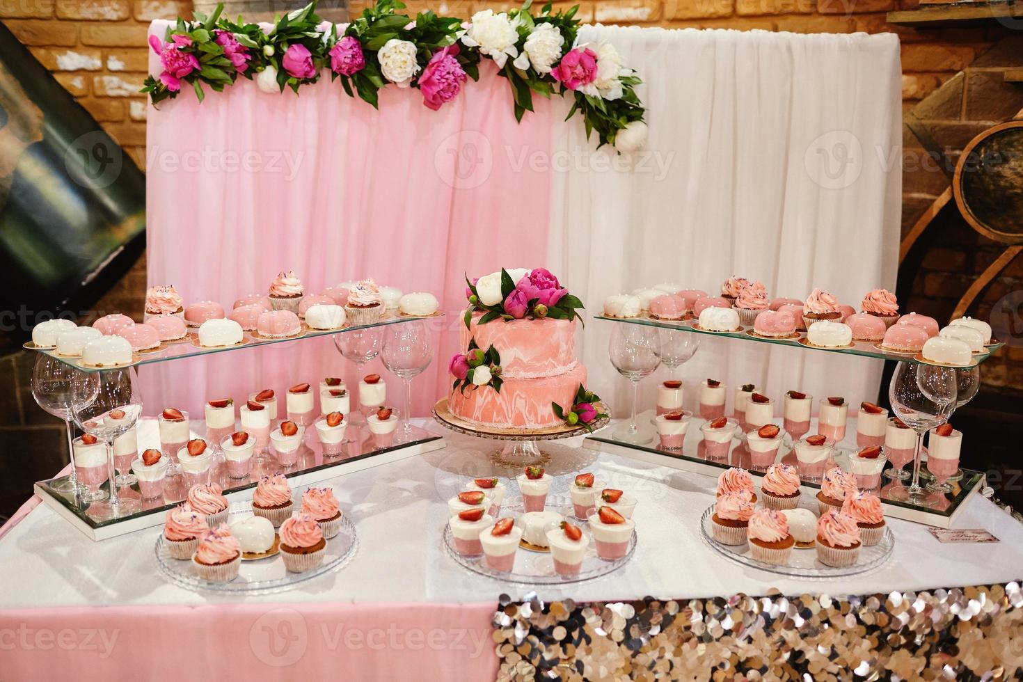 rosa Hochzeitstorten des Schokoriegels, die durch Blumen verziert werden, die an einem festlichen Tisch mit Wüsten, Erdbeertörtchen und Cupcakes stehen. Hochzeitskonzept foto