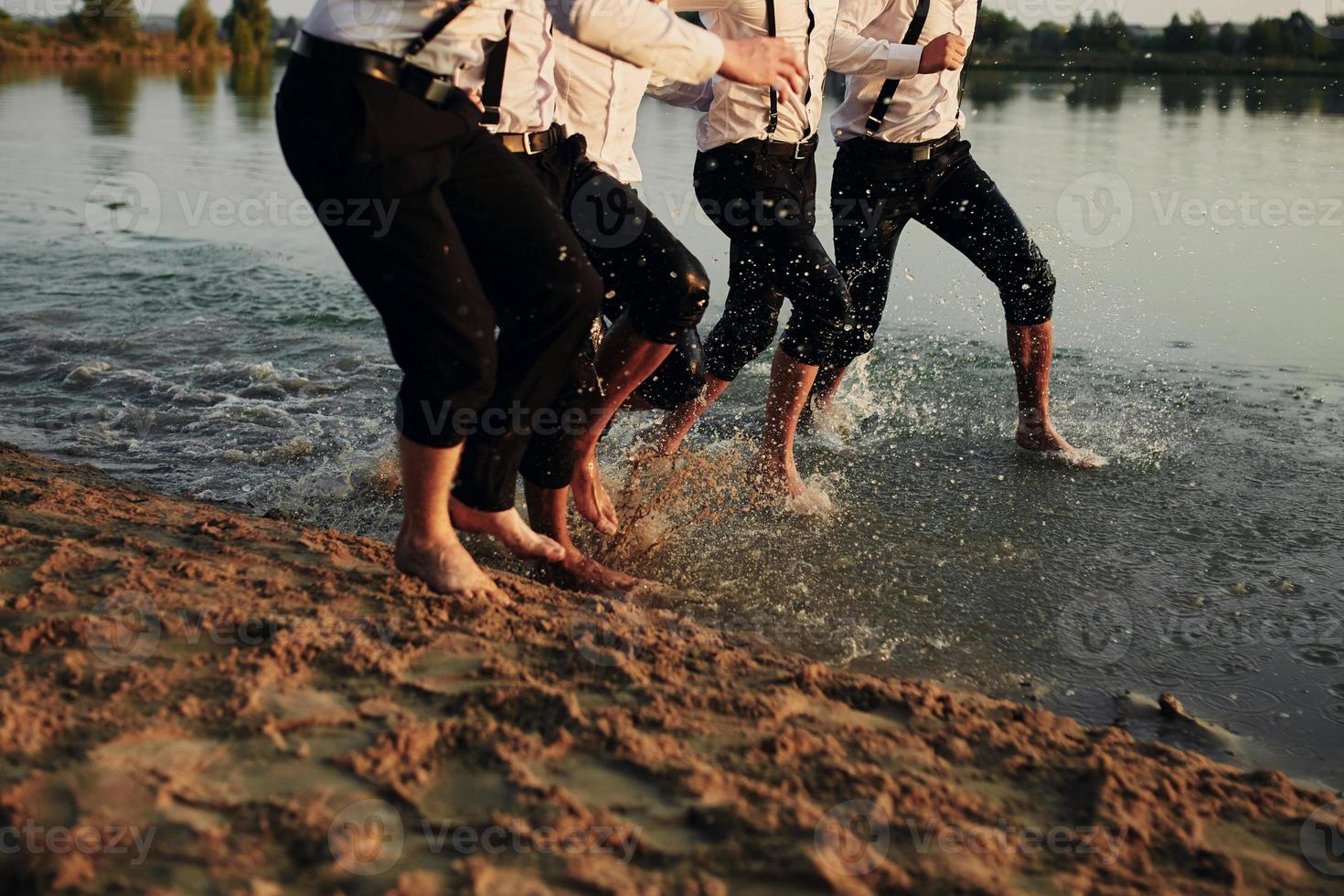 Männerfüße im Wasser. Männer in Kostümen laufen auf dem Wasser. Sie haben Spaß, spielen und spritzen Wasser um sich herum. Sommer. Eine Gruppe glücklicher junger Männerfüße spritzt Wasser ins Meer und sprüht am Strand foto