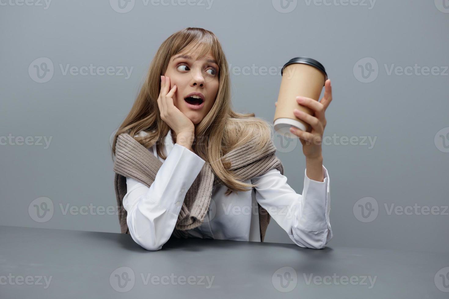 schockiert jung blond Schüler Dame im warm Sweatshirt aussehen beim wegbringen Kaffee schließen Mund Sitzung beim Arbeitsplatz im grau modern Zuhause Büro. Kaffee brechen Liebhaber Konzept. Kopieren Raum. cool Angebot foto