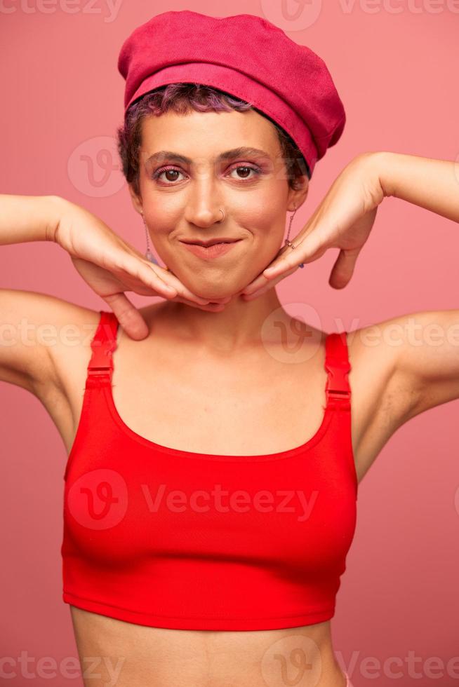 jung sportlich Frau mit ein kurz Haarschnitt und lila Haar im ein rot oben und ein Rosa Hut mit ein sportlich Zahl lächelt und Grimassen suchen beim das Kamera auf ein Rosa Hintergrund foto