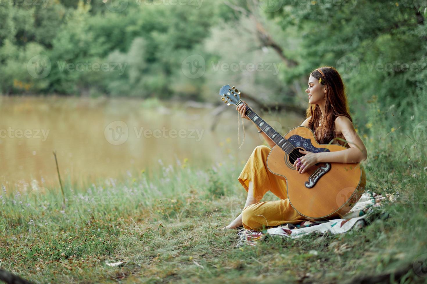 Hippie Frau spielen Gitarre lächelnd und Singen Lieder im Natur Sitzung auf ein Plaid durch das See im das Abend im das Strahlen von das Rahmen Sonne. ein Lebensstil im Harmonie mit das Körper und Natur foto