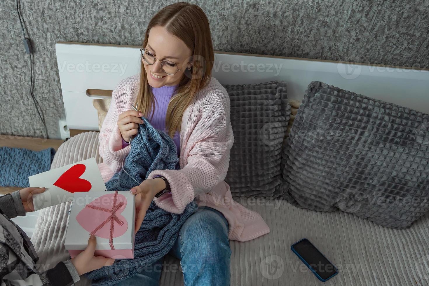 das Kind gratuliert seine Mutter auf Mutter Tag, gibt ihr ein Box mit ein Geschenk und ein Glückwunsch Öffner mit ein Herz gemacht durch sich selbst. ein Frau strickt im ihr Schlafzimmer. foto