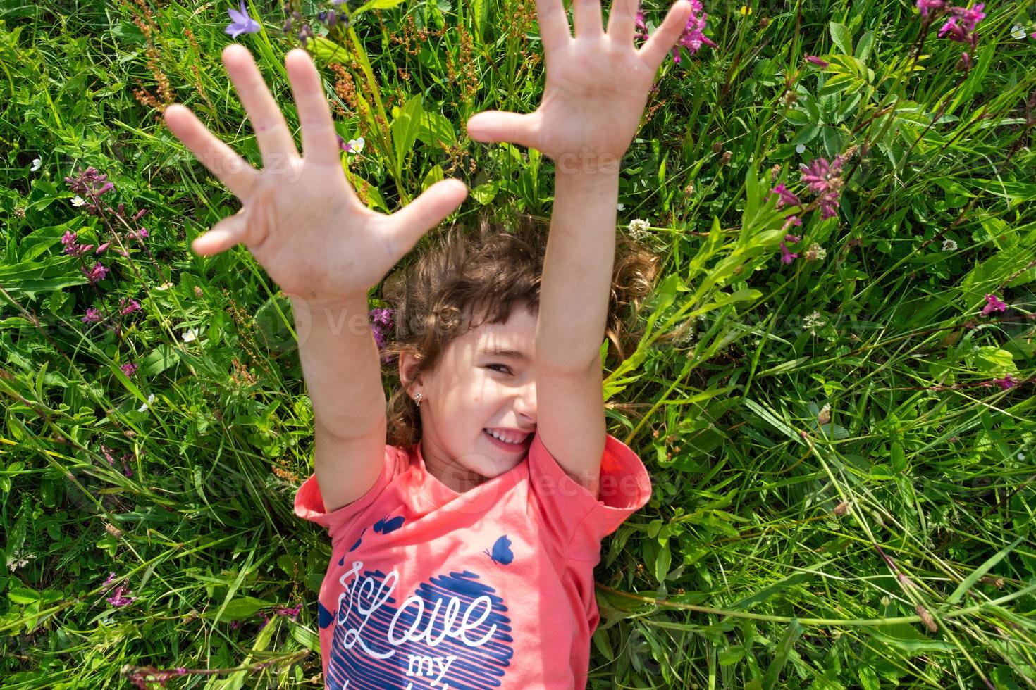 Porträt von ein Kind Mädchen im Sommer- Lügen im das Gras und Wildblumen mit Absätze und Palmen. Sommer- Zeit, Freiheit foto