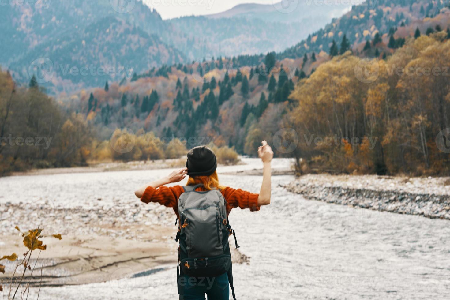 ein Reisender im ein Sweatshirt Hut mit ein Rucksack Gesten mit ihr Hände und entspannt sich auf das Fluss Bank im das Berge foto