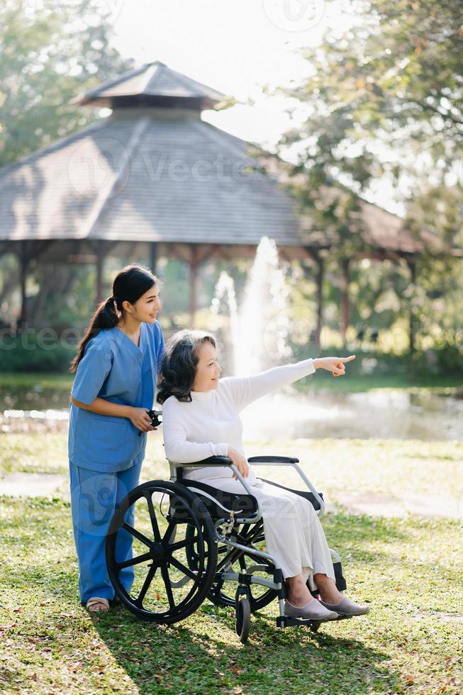 Alten asiatisch Senior Frau auf Rollstuhl mit asiatisch Vorsichtig Betreuer. Pflege- Zuhause Krankenhaus Garten Konzept. im Sonnenlicht foto