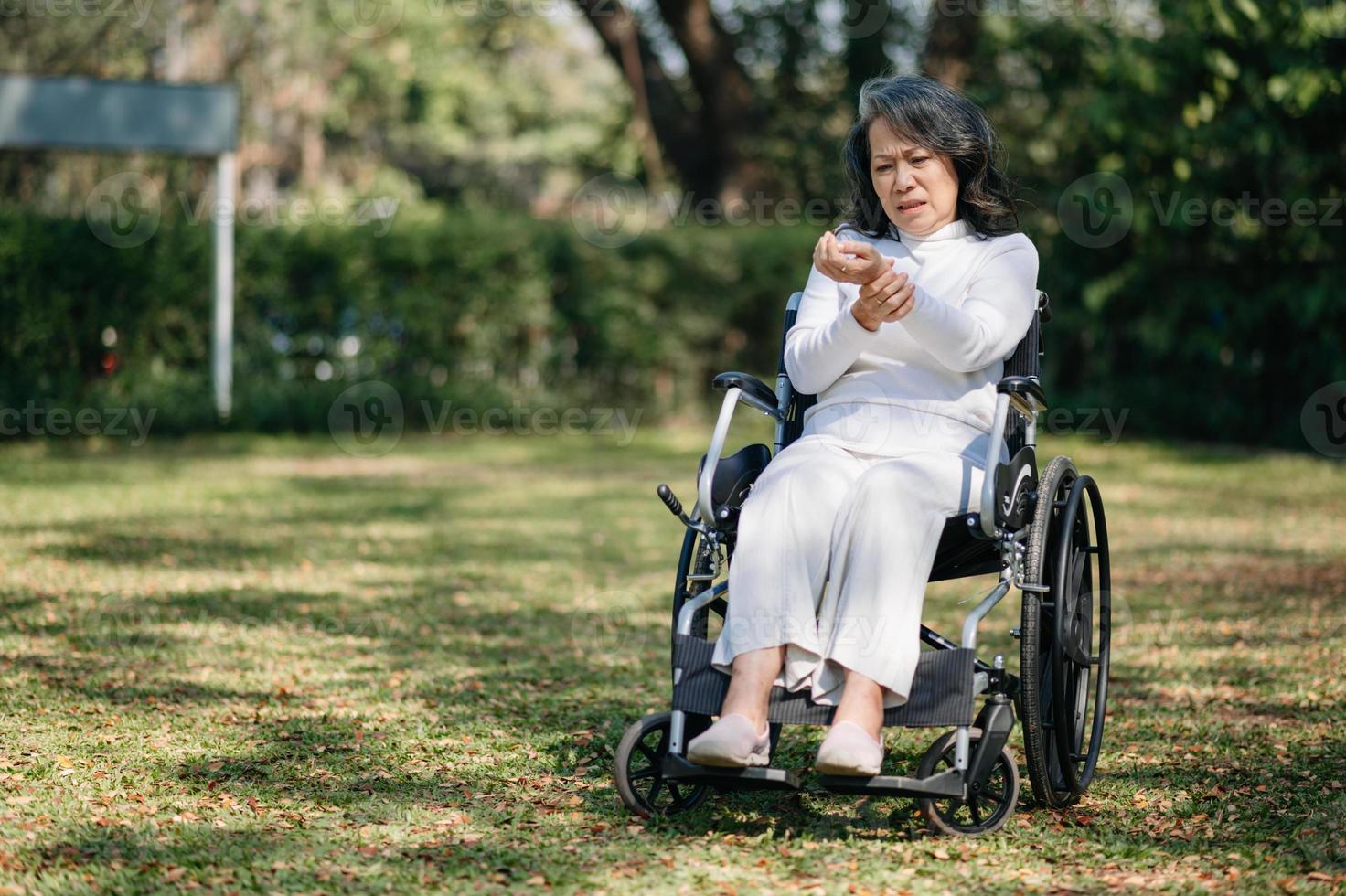 asiatisch alt Frau Sitzung auf ein Rollstuhl draußen im das Park haben Schmerzen im das Waffen, Handgelenke und Körper im Sonne Licht foto