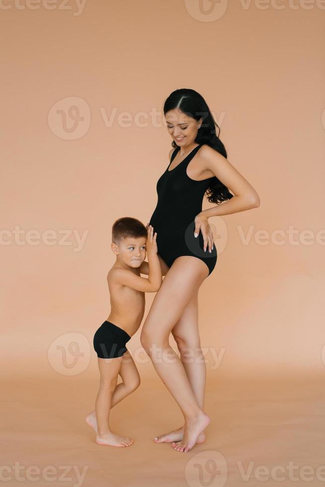 Porträt von ein schön glücklich schwanger Frau und ihr süß Baby, isoliert Bild auf ein Beige Hintergrund. Sohn Umarmungen Mamas Bauch foto