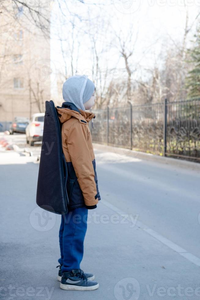 ein Teenager Junge im ein Jacke und Hut Spaziergänge allein mit ein Gitarre im ein Fall. Kind Musiker hat verloren im das Stadt foto