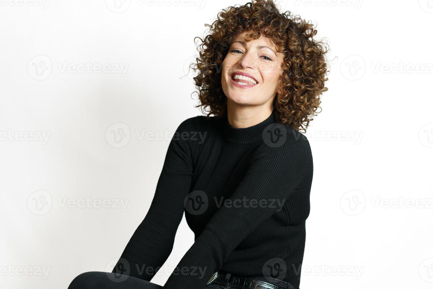 lächelnd Frau mit lockig Haar Sitzung gegen Weiß Mauer foto