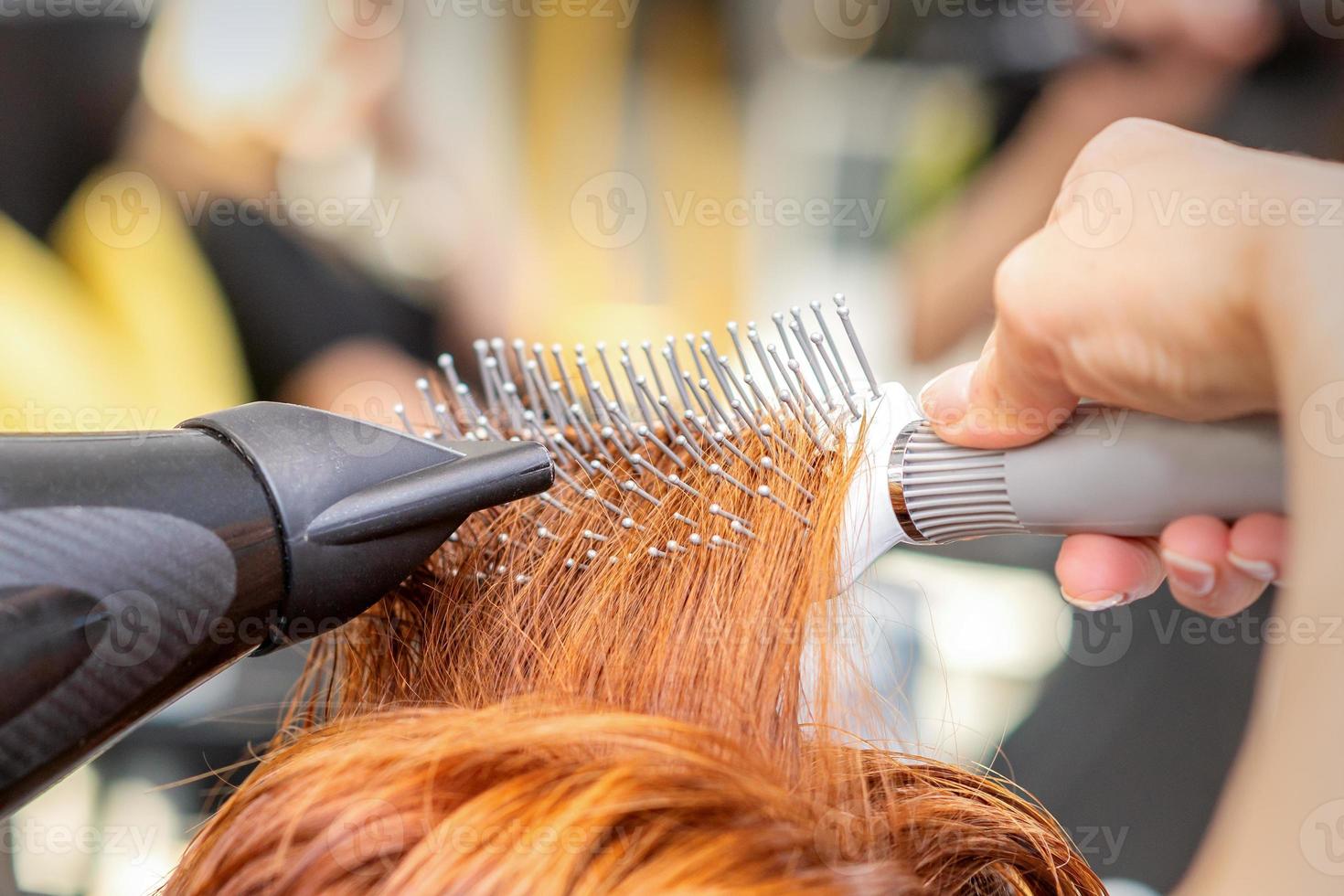 Nahansicht von Meister Hand mit Föhnen und Haarbürste weht weiblich rot Haar im ein Salon. foto