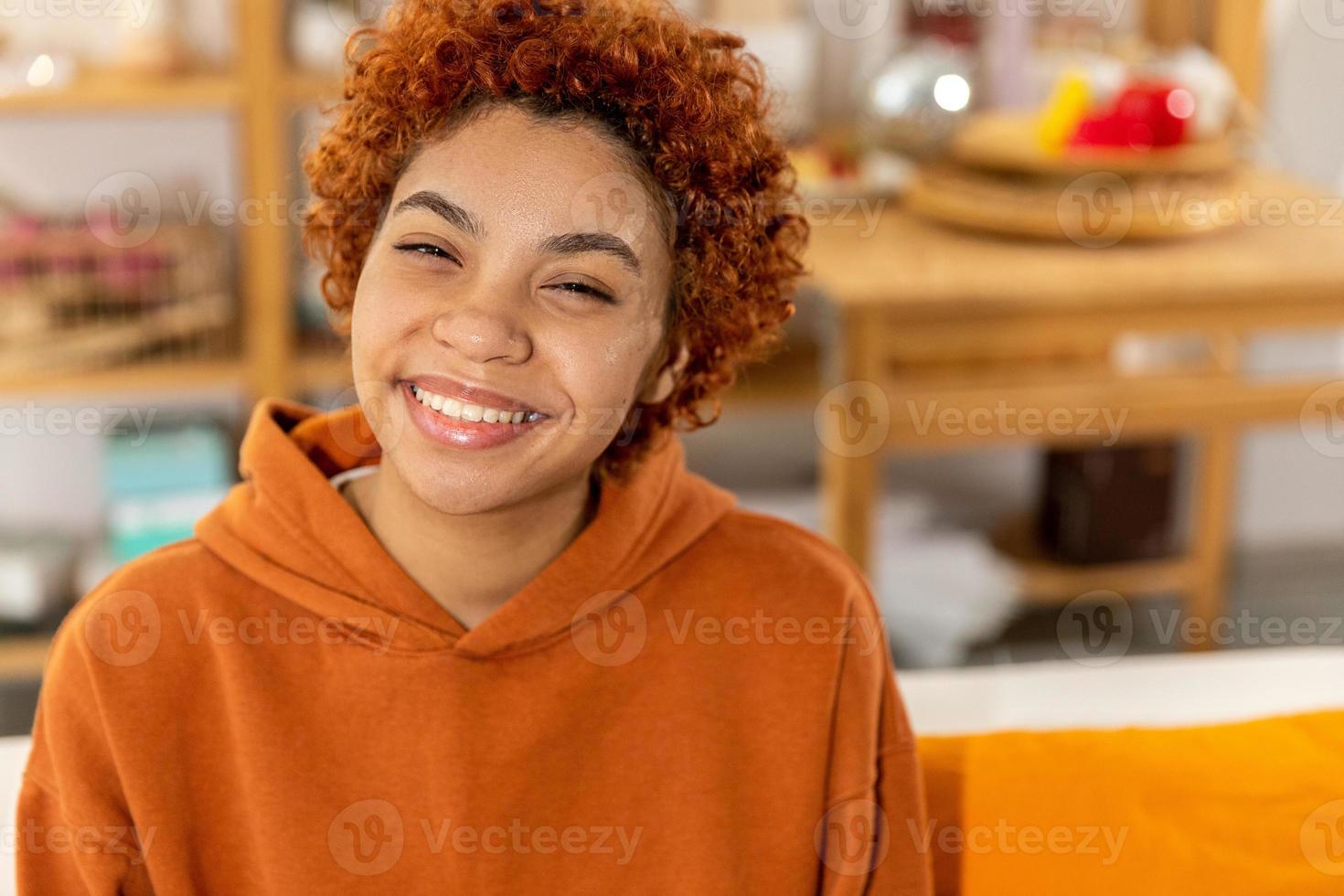 Schönes afrikanisch-amerikanisches Mädchen mit Afro-Frisur lächelnd auf dem Sofa zu Hause drinnen sitzend. Junge afrikanische Frau mit lockigem Haar lacht. freiheit glück sorglos glückliche menschen konzept. foto