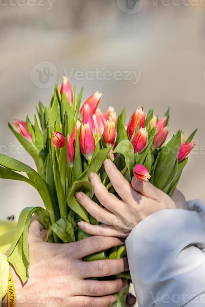 ein jung Paar im Liebe auf das Straße, halten Blumen im ihr Hände beim ein romantisch Moment. ein Mann gibt rot Tulpen zu ein blond Frau.die Konzept von das Frühling Urlaub ist März 8, Ostern, Damen Tag foto