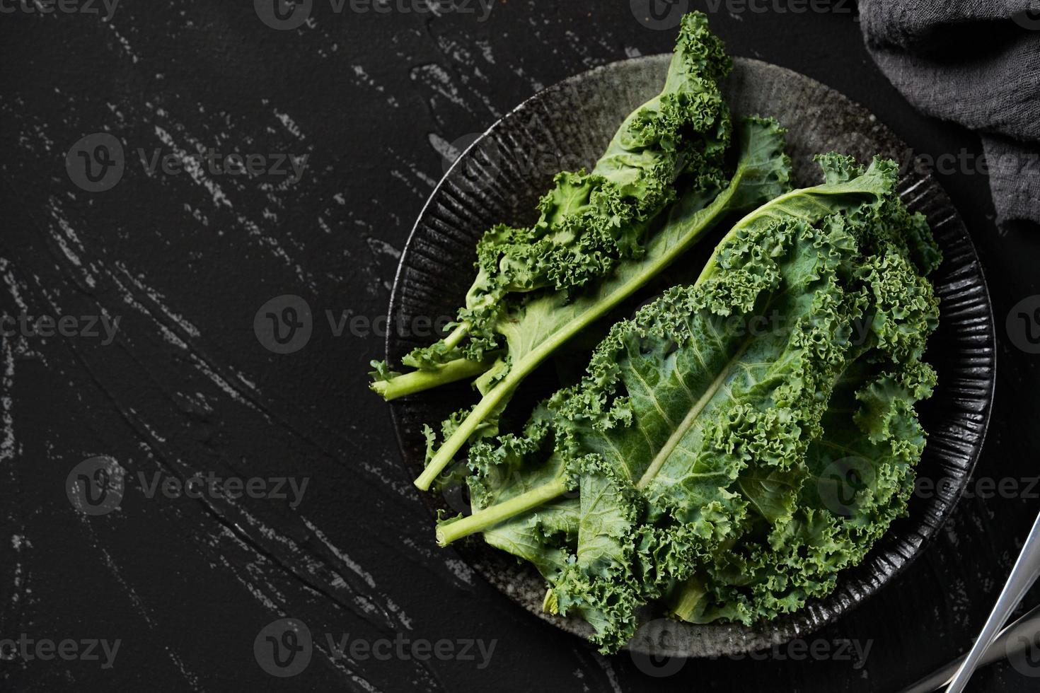 Konzept von frisch Grünkohl Blätter Salat im ein schwarz Gericht auf dunkel Hintergrund. Grün Grünkohl Blätter Salat Essen im das Küche. eben legen, oben Sicht, Löffel, Gabel foto