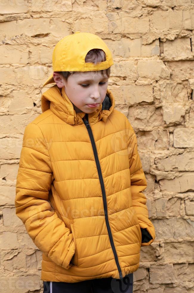 ein Kind auf das Straße. ein Übergewicht Junge ist Stehen gegen ein Backstein Mauer im Gelb Kleider und ein Baseball Deckel. foto