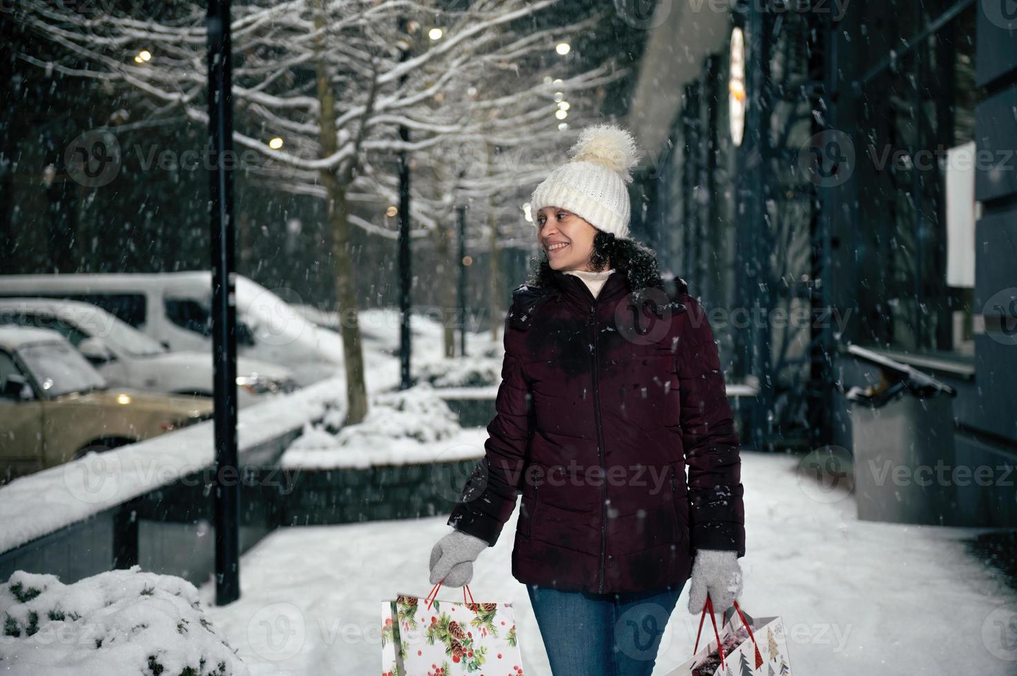 herrlich multiethnisch Frau im warm Kleidung, mit Einkaufen Taschen, Gehen Nieder das Stadt Straße, auf ein schneebedeckt Winter Nacht foto