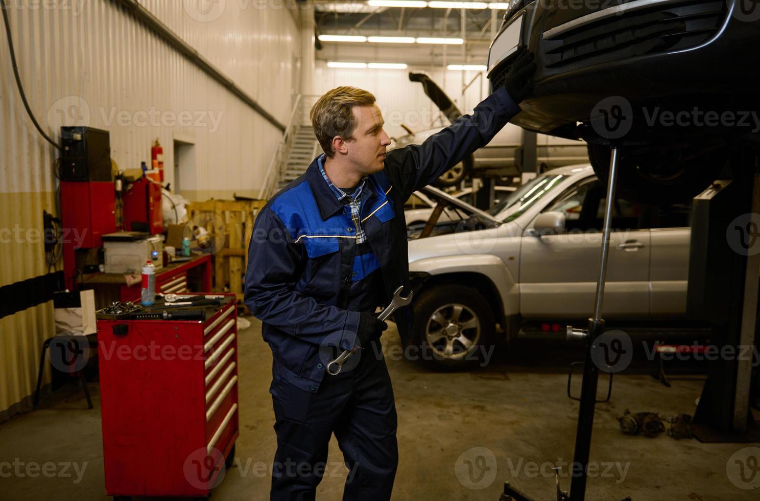 gut aussehend jung Mechaniker im Fachmann Uniform, halten ein Schlüssel und Arbeiten im Auto Bedienung mit angehoben Fahrzeug. Auto Reparatur und Wartung. foto