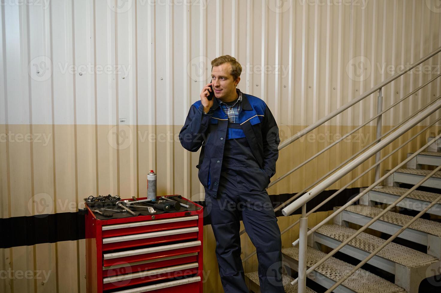 Porträt von ein gut aussehend kaukasisch Mann, Techniker, Garage Mechaniker reden auf Handy, Mobiltelefon Telefon Stehen in der Nähe von ein Box mit Arbeit Werkzeuge zum prüfen und Reparatur Autos und Auto Bedienung Bahnhof foto