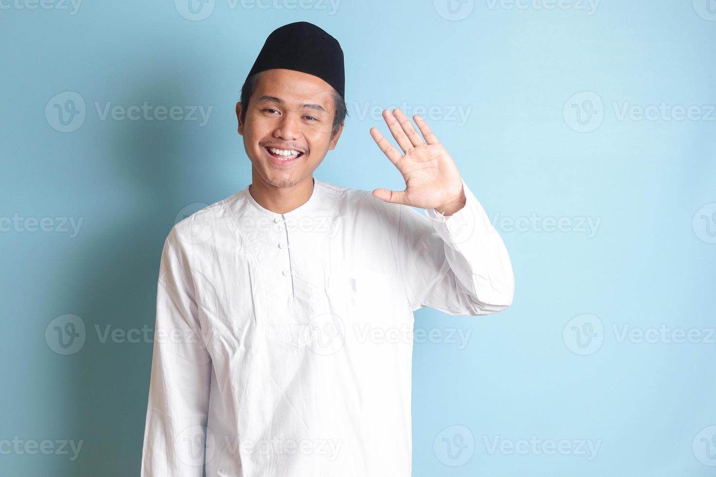 Porträt von asiatisch Muslim Mann im Weiß koko Hemd mit Schädeldecke Hemd Sprichwort Hi, winken Hand beim Kamera, Gruß Person mit glücklich lächeln. isoliert Bild auf Blau Hintergrund foto