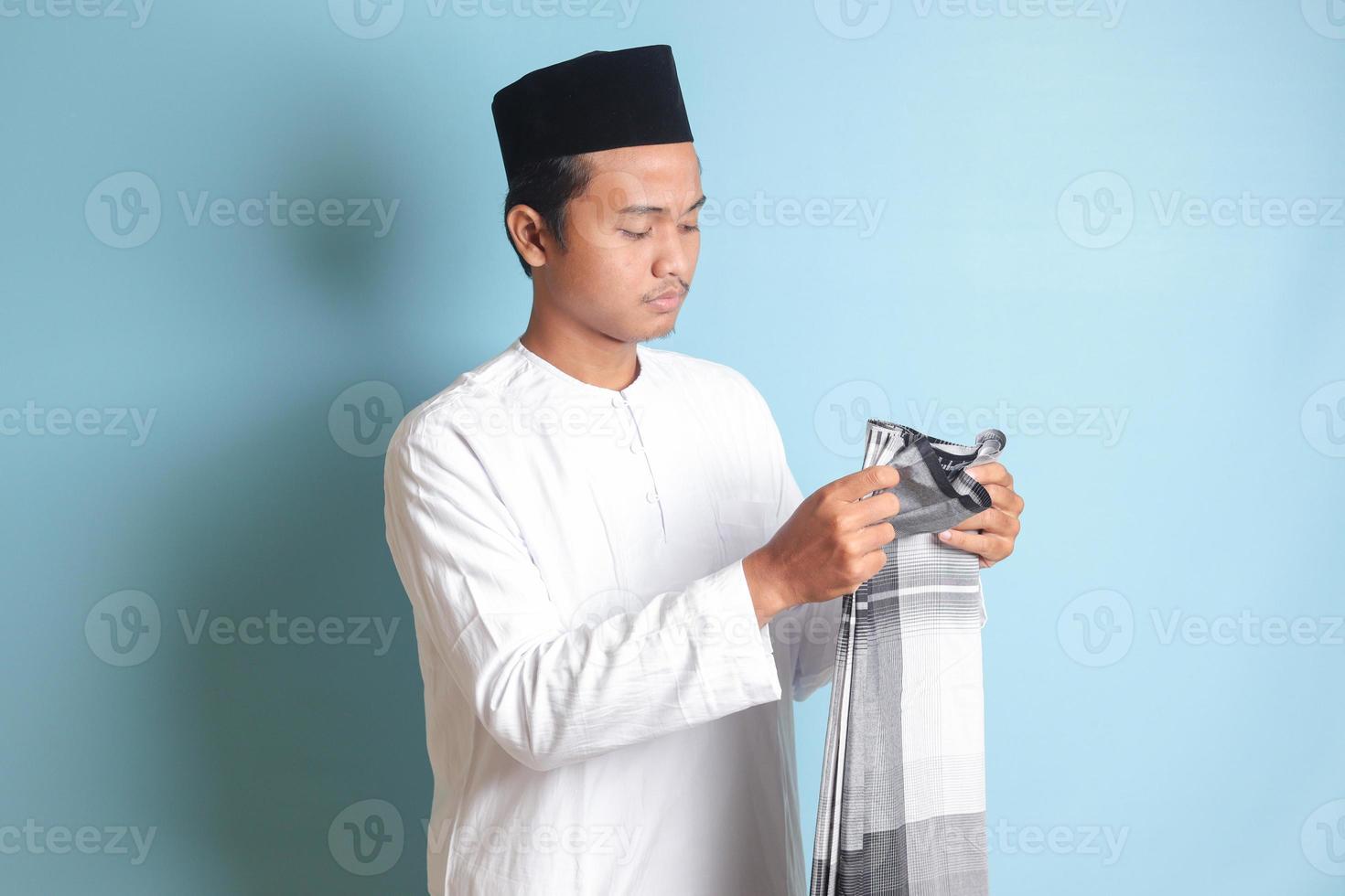 Porträt von asiatisch Muslim Mann im Weiß koko Hemd falten und aufgeräumt oben seine Sarung . isoliert Bild auf Blau Hintergrund foto