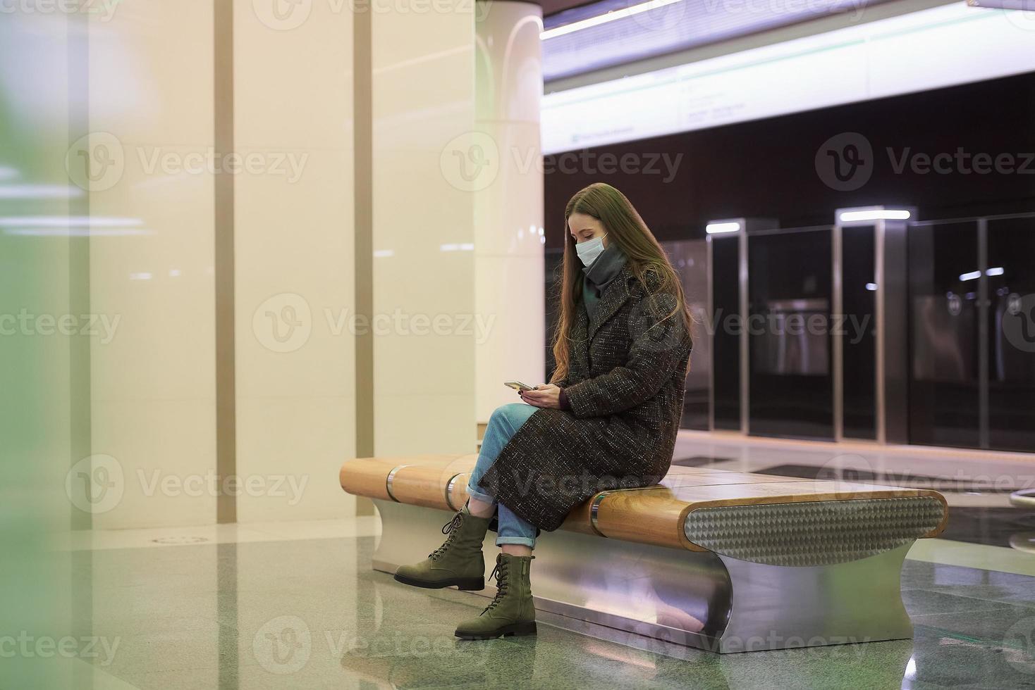 Eine Frau in einer medizinischen Gesichtsmaske wartet auf einen Zug und hält ein Smartphone in der Hand foto