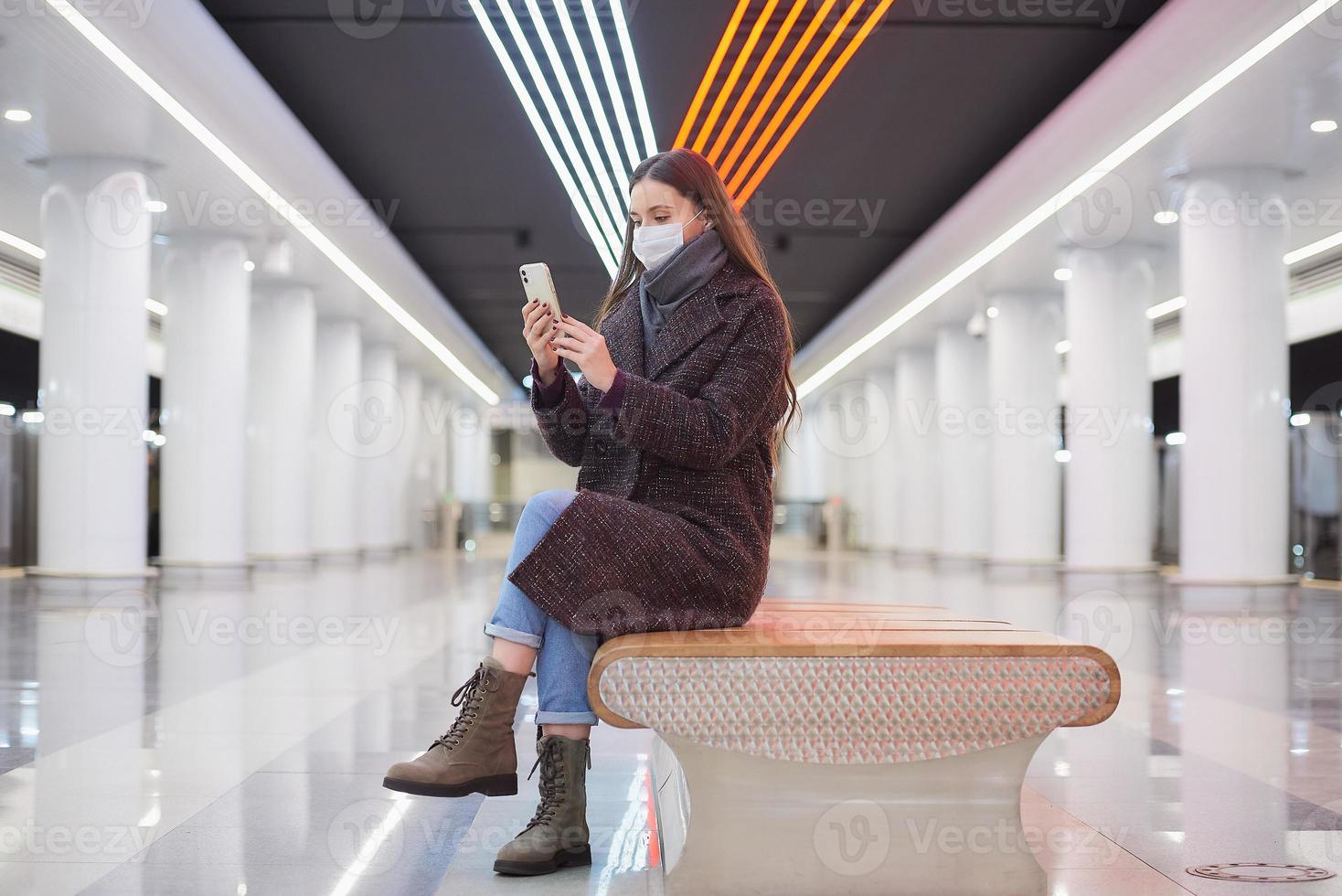 Eine Frau in einer medizinischen Gesichtsmaske wartet auf einen Zug und hält ein Smartphone in der Hand foto