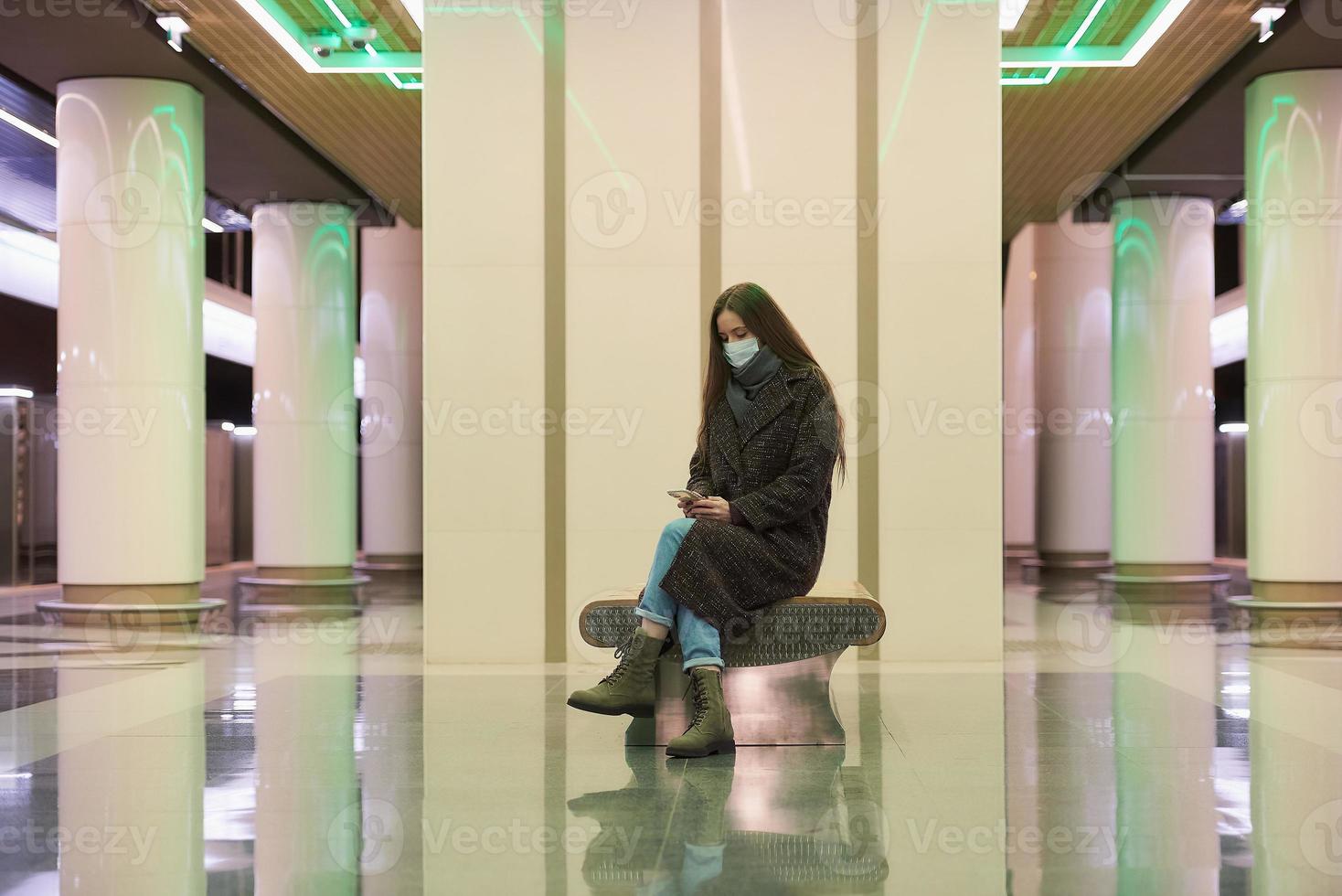 Eine Frau in einer medizinischen Gesichtsmaske wartet auf einen Zug und hält ein Smartphone in der Hand foto