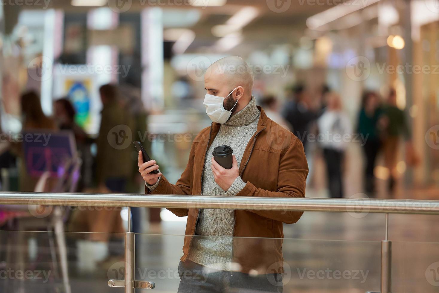Mann in einer Gesichtsmaske benutzt ein Telefon und hält einen Kaffee im Einkaufszentrum foto