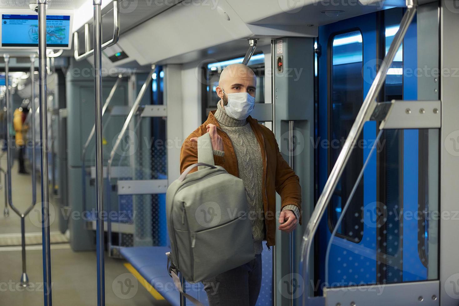 Ein kahlköpfiger Mann mit Bart in einer Gesichtsmaske zieht in einem U-Bahn-Wagen einen Rucksack an foto