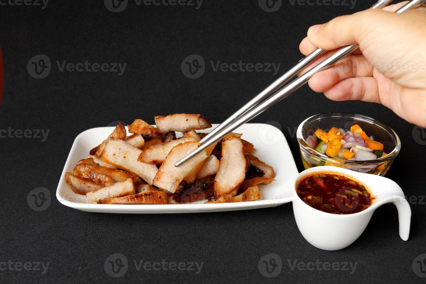 richtig Hand verwenden Essstäbchen klemmen gegrillt Schweinefleisch Schulter Scheibe im Weiß Rechteck Gericht mit heiß und sauer thailändisch Stil Soße im Weiß Tasse und Gemüse Scheibe im Essig Glas Tasse auf schwarz Hintergrund. foto