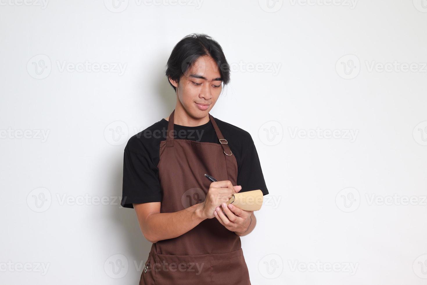 Porträt von attraktiv asiatisch Barista Mann im braun Schürze nehmen Befehl, Schreiben auf Speisekarte Buch Liste. isoliert Bild auf Weiß Hintergrund foto