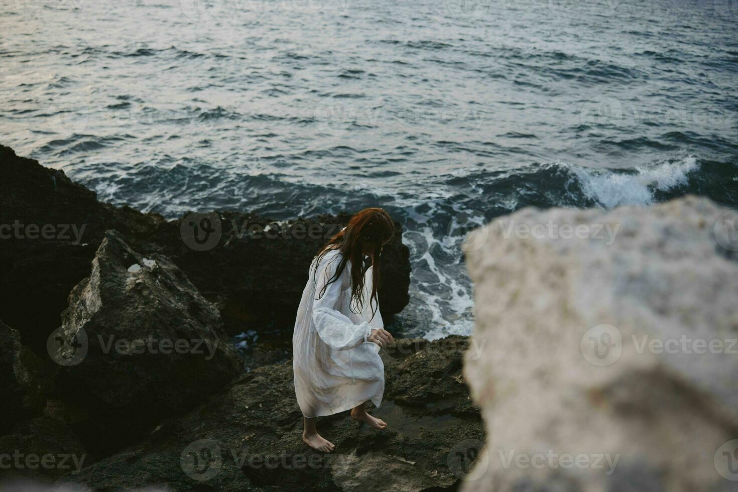 ein Frau im ein Weiß Kleid steht barfuß auf das Felsen durch das Ozean foto
