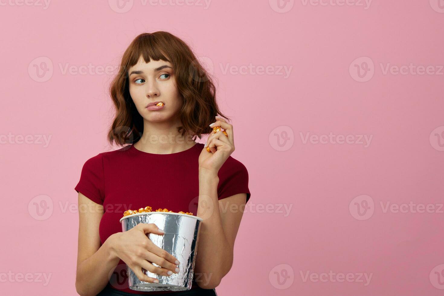 aufgeregt verwirrt süß Rothaarige Dame im rot T-Shirt mit Popcorn bereit zum Film Abend posieren isoliert auf Über Rosa Studio Hintergrund. Kopieren Raum Banner. Mode Kino Konzept. Unterhaltung Angebot foto