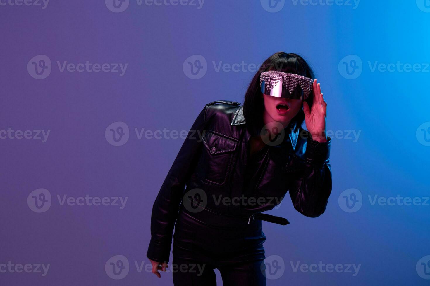 aufgeregt ziemlich genial brünett Frau im Leder Jacke berühren modisch spiegelnd Sonnenbrille öffnen Mund posieren isoliert im Blau violett Farbe Licht Hintergrund. Neon- Party Cyberpunk Konzept. Kopieren Raum foto