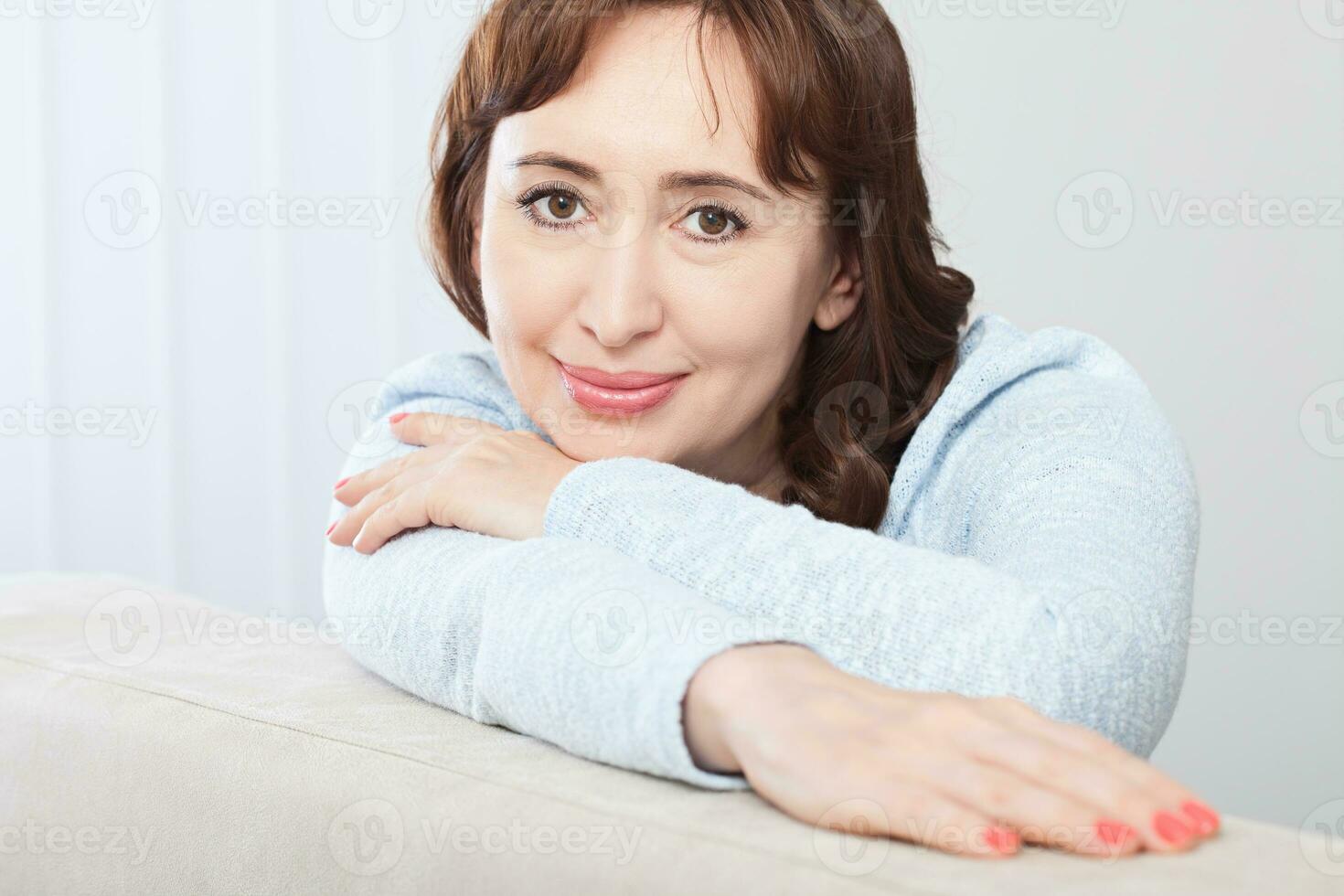 schön mittleren Alters Brünette Frau mit ein strahlend Lächeln Sitzung auf ein Sofa beim Zuhause suchen beim das Kamera foto